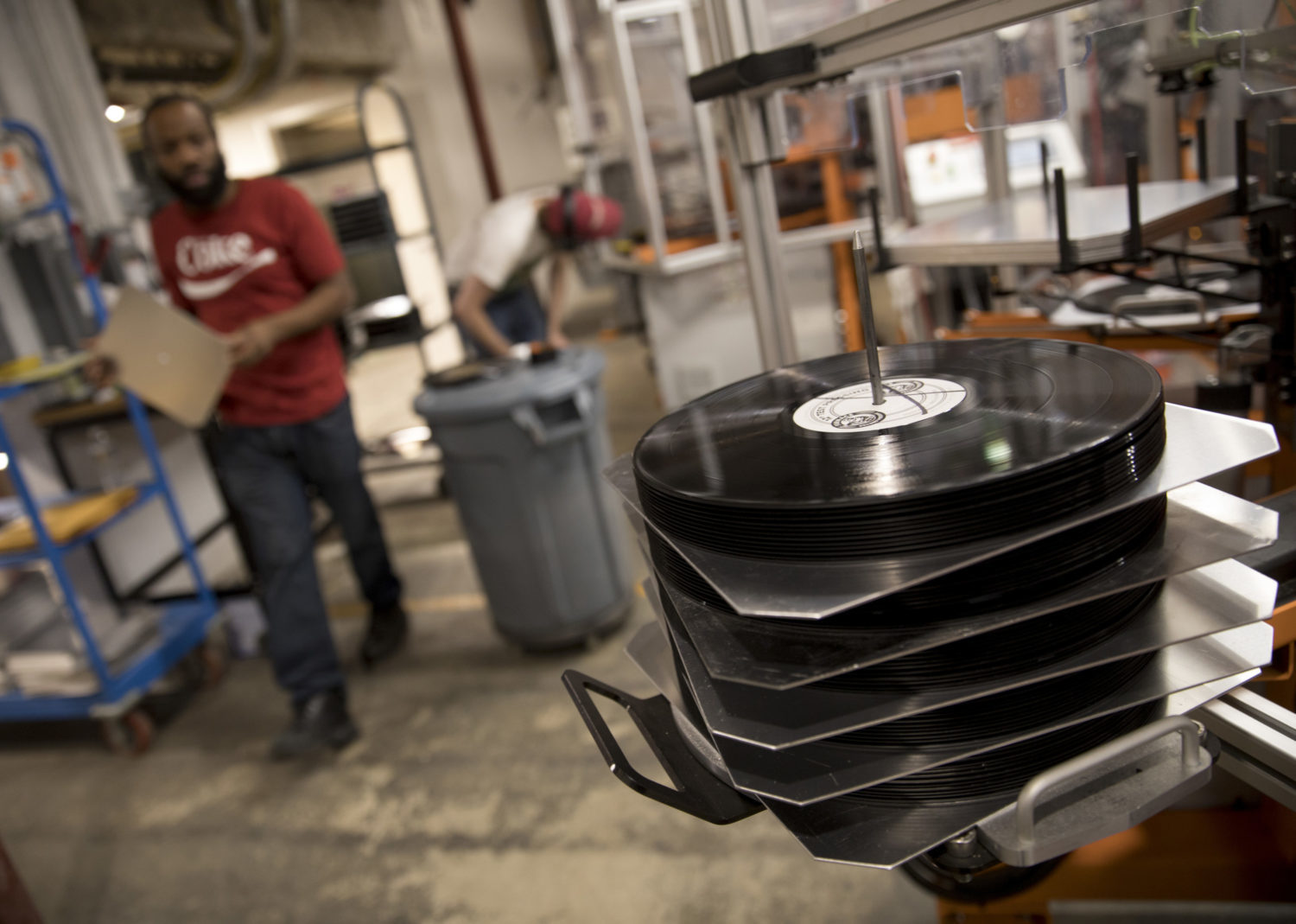 Several manufactured records are automatically stacked at a pressing plant while an employee walks in the background.